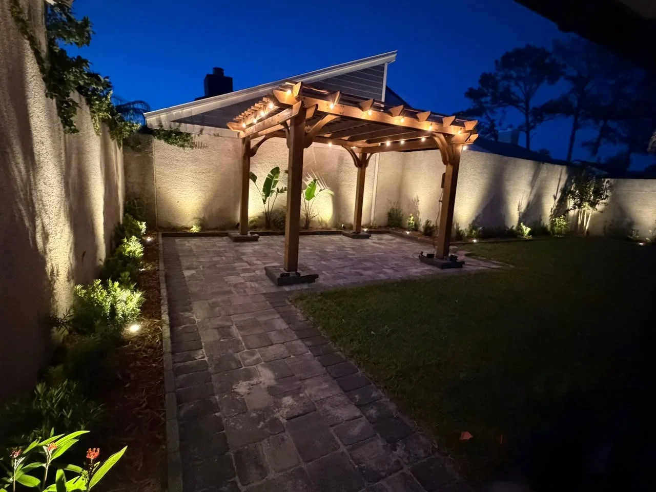 Wooden pergola with string lights over paver patio, illuminated concrete walls, and landscape lighting